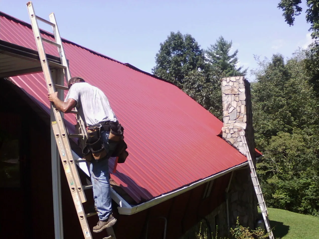Roofer on a ladder beside a red metal roof and stone chimney — Soffit & Fascia Repair by Roofing Brothers in Pawleys Island